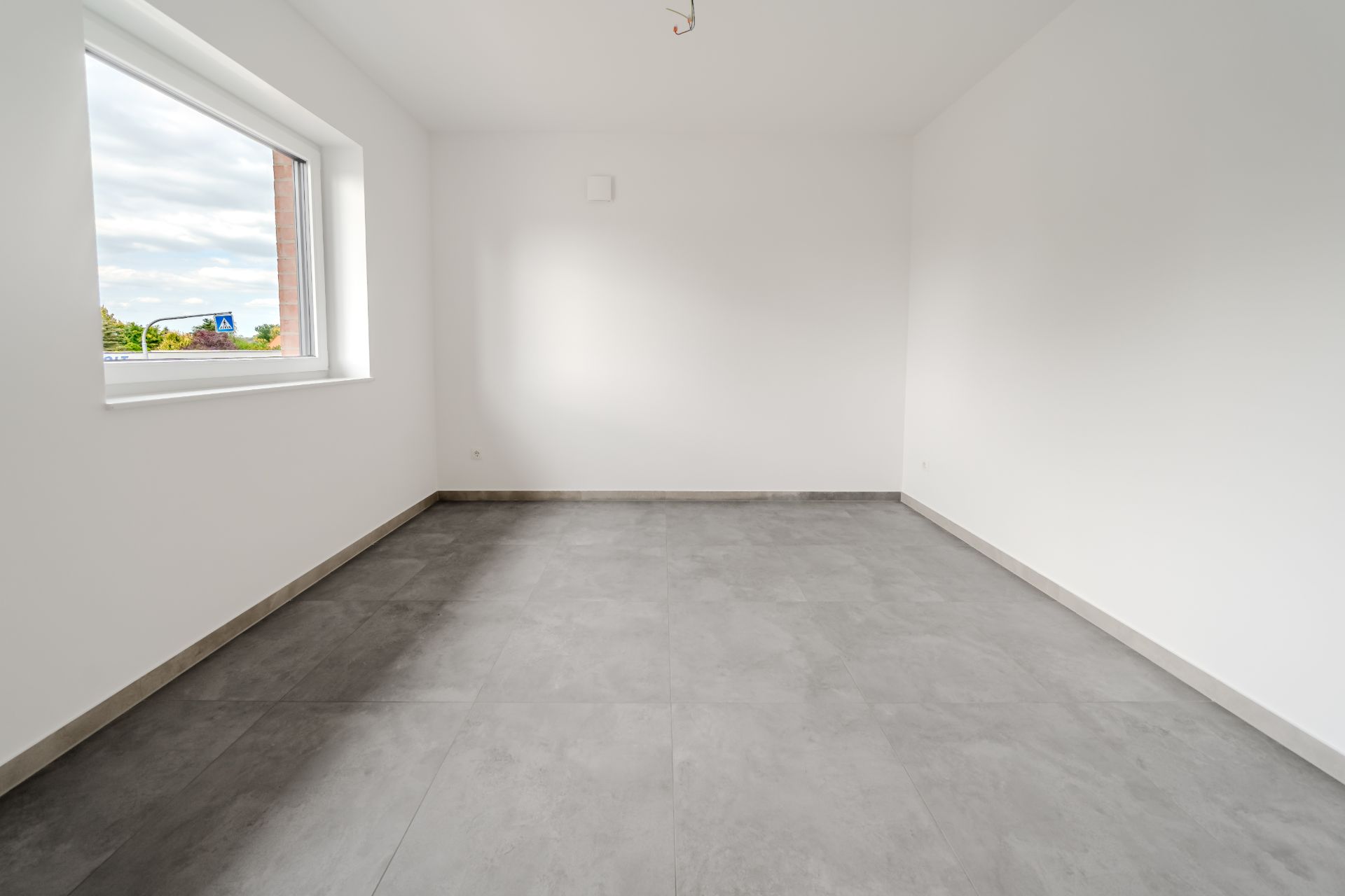 Leerer Raum mit grauem Fliesenboden, weißen Wänden und großem Fenster.