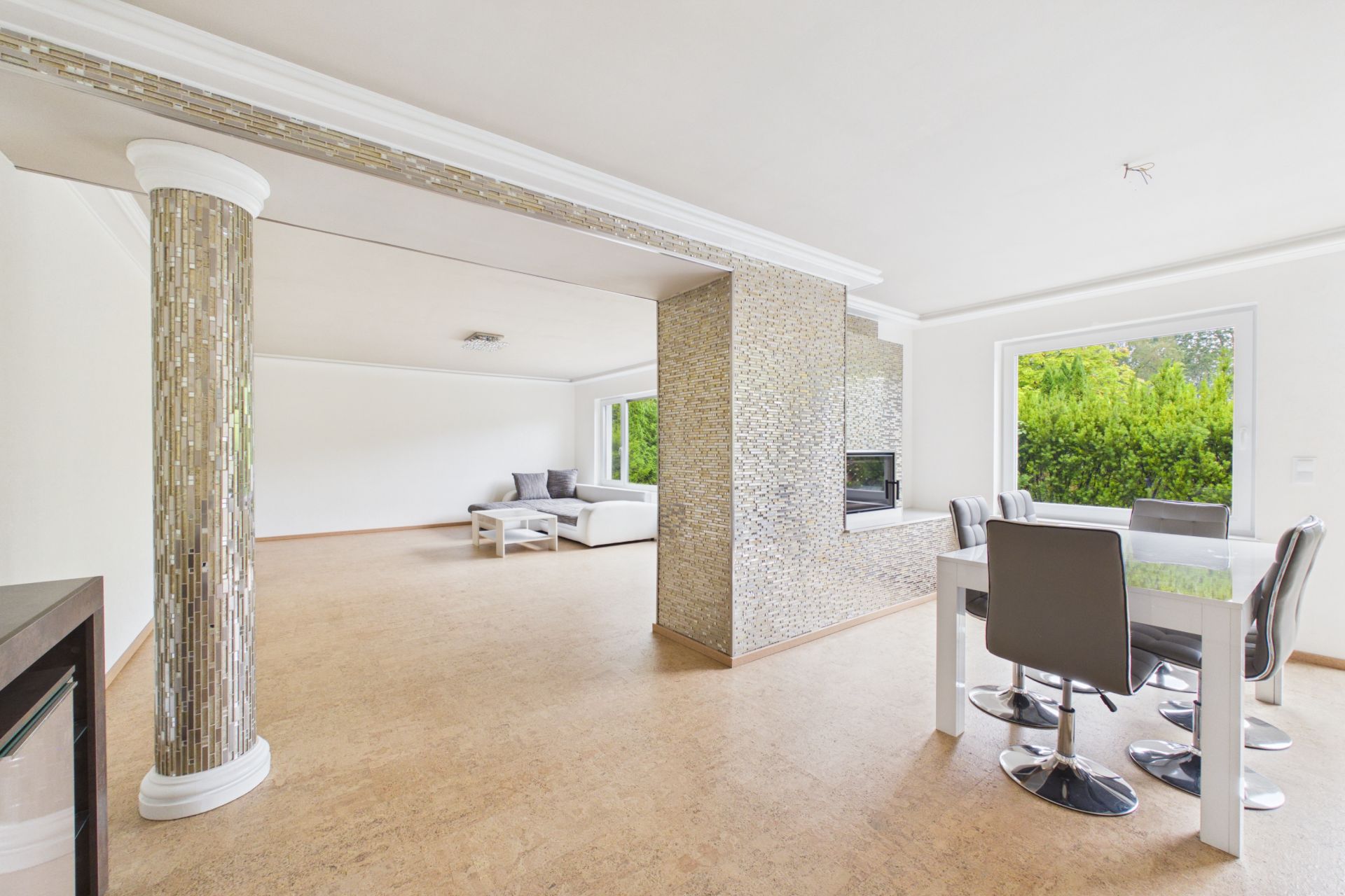 Helles Wohnzimmer mit Glastisch, Stühlen, Sofa und verzierten Säulen, große Fenster mit Blick ins Grüne.