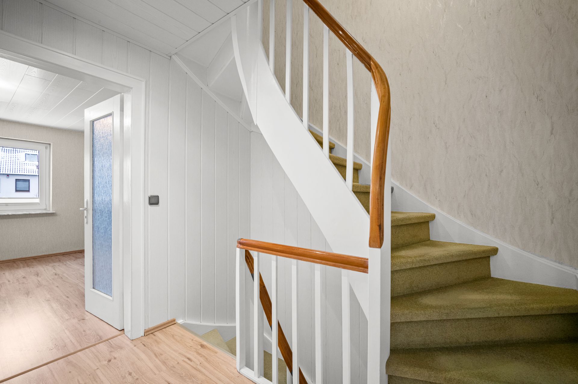 Innenraum mit Treppenhaus, weißen Wänden, offener Tür und Fenster im Hintergrund.