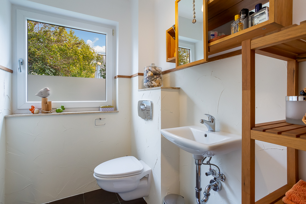 Kleines Badezimmer mit Toilette, Waschbecken, Fenster und Holzregal.