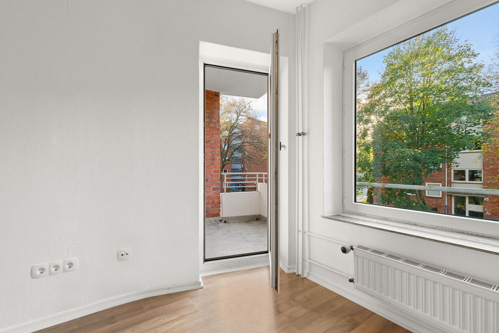 Helles Zimmer mit Holzboden, großem Fenster und Zugang zu einem Balkon.