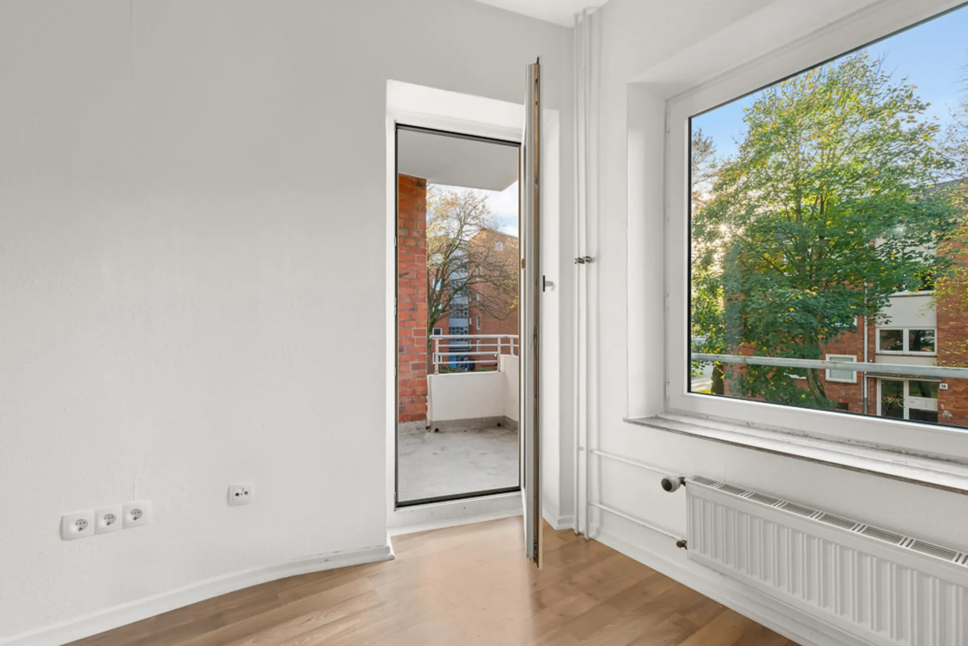 Helles Zimmer mit Holzboden, großem Fenster und Zugang zu einem Balkon.