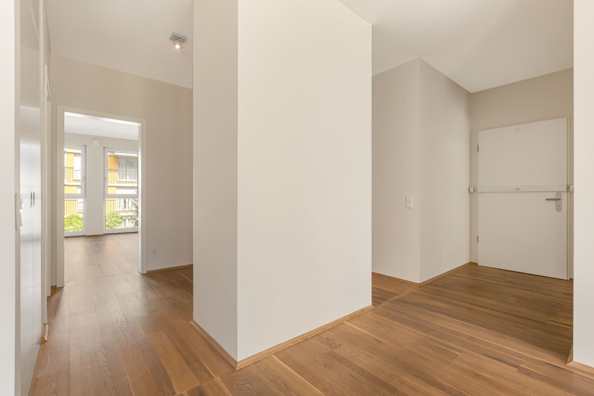 Heller Flur mit Holzboden, weißer Wand und Tür, Zugang zu einem Raum mit großen Fenstern.