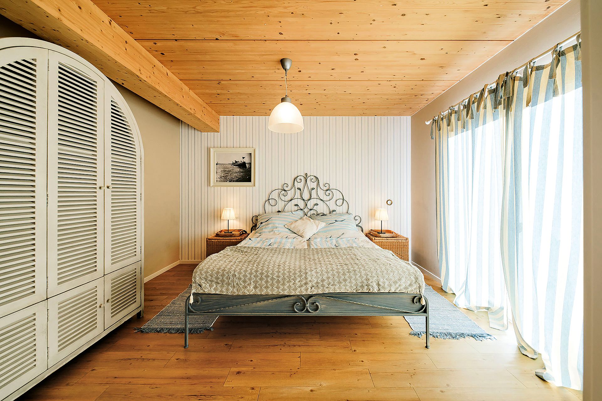 Helles Schlafzimmer mit Doppelbett, großem Kleiderschrank und bodentiefen Fenstern mit Vorhängen.