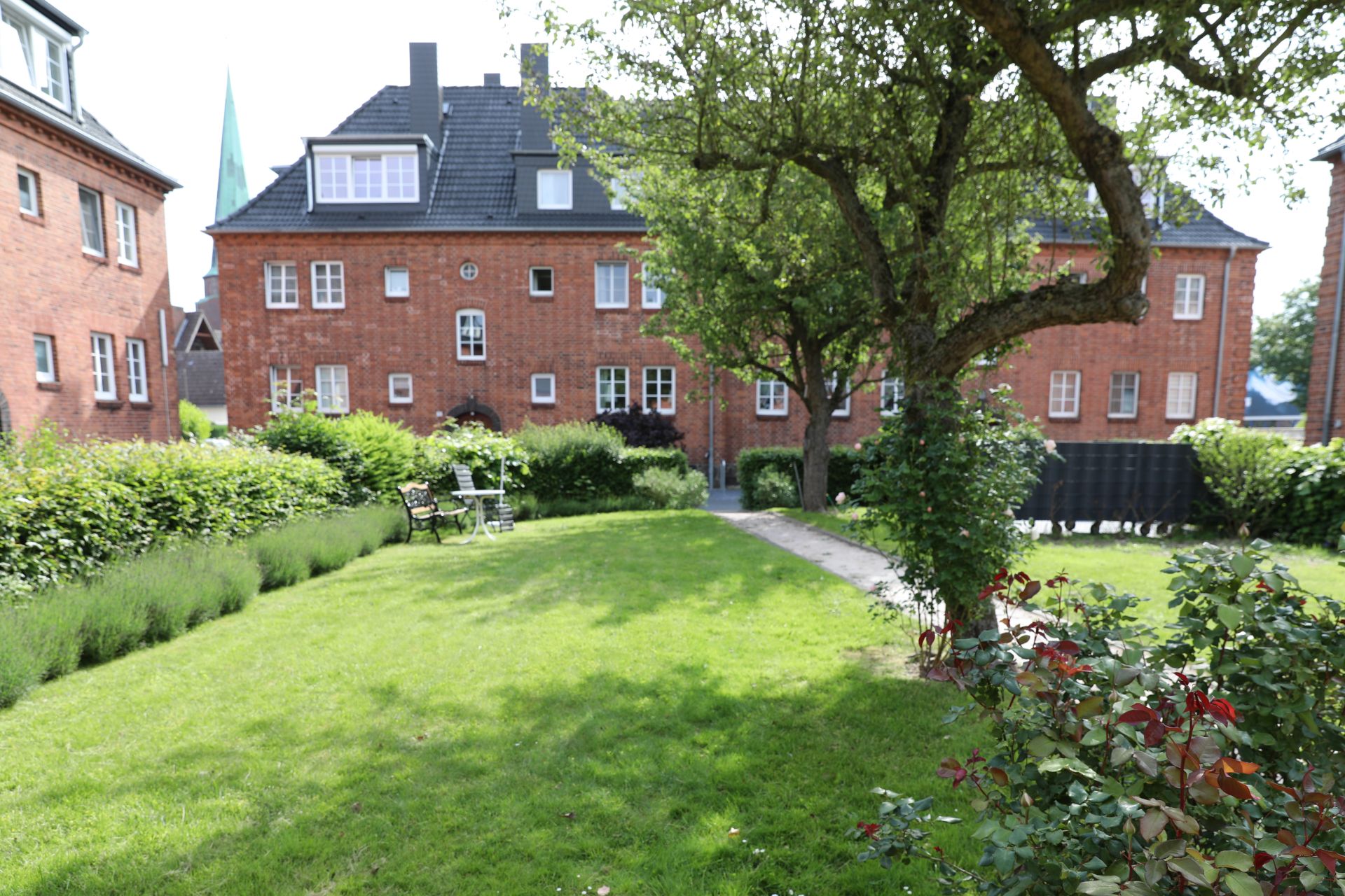 Grüner Garten mit Bäumen, Sträuchern, Rasenfläche und Sitzbank vor roten Backsteinhäusern.