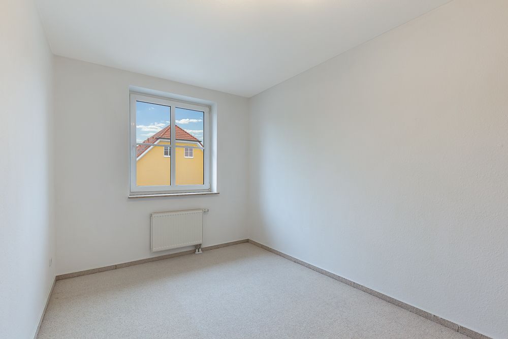 Leerer, heller Raum mit weißem Wänden, Teppichboden und Fenster mit Blick auf ein gelbes Haus.
