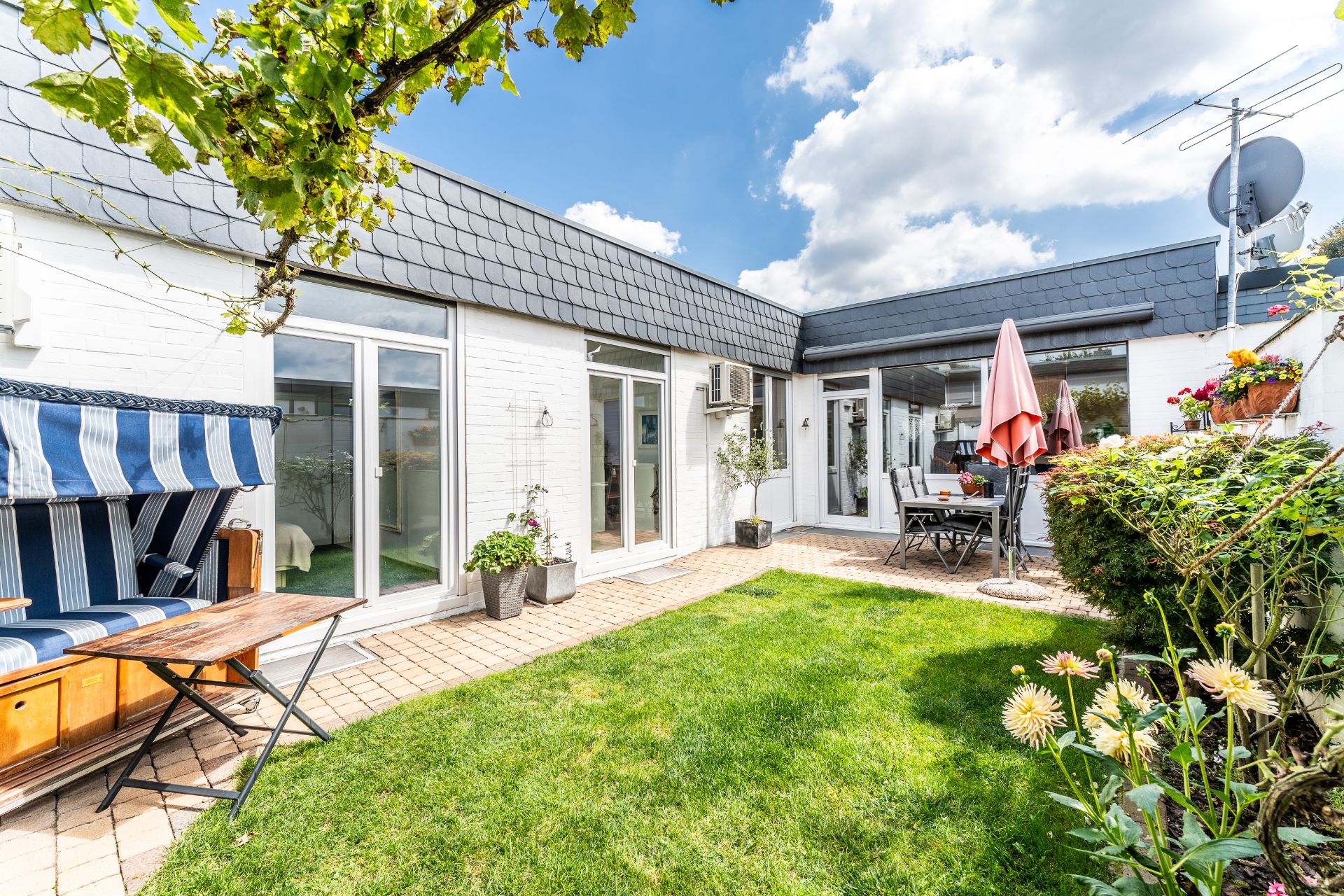 Grüner Innenhof mit Gartenmöbeln, Strandkorb, Blumen und Terrassentüren bei sonnigem Wetter.