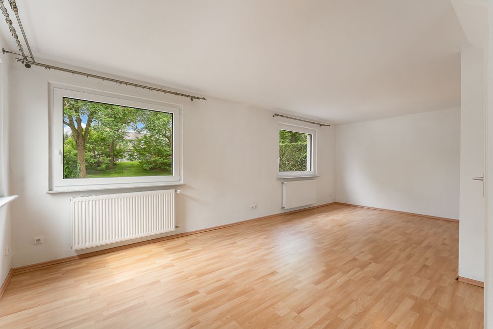 Leerer Raum mit hellem Holzboden, weißen Wänden und zwei großen Fenstern mit Blick ins Grüne.