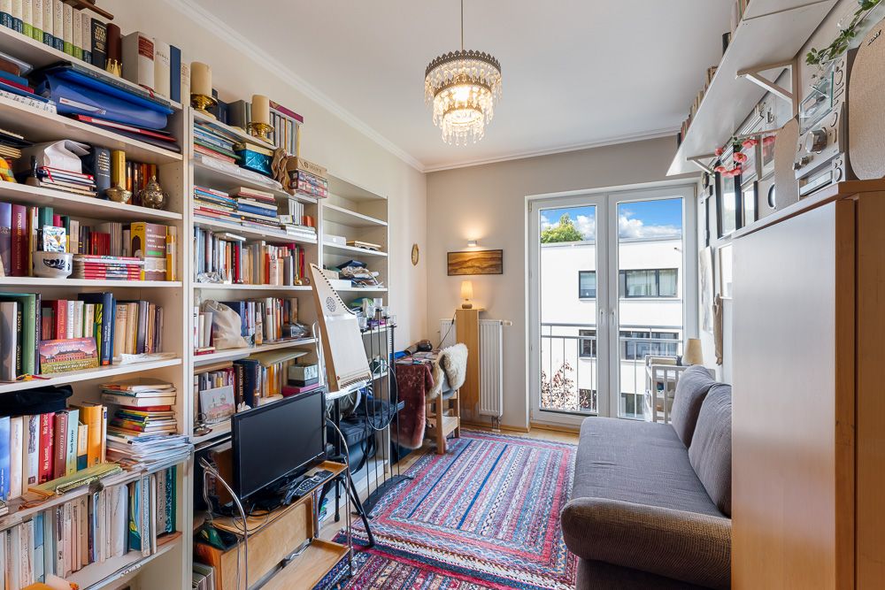 Wohnzimmer mit Sofa, Bücherregalen, Schreibtisch, Teppich und Balkon, viel Tageslicht.