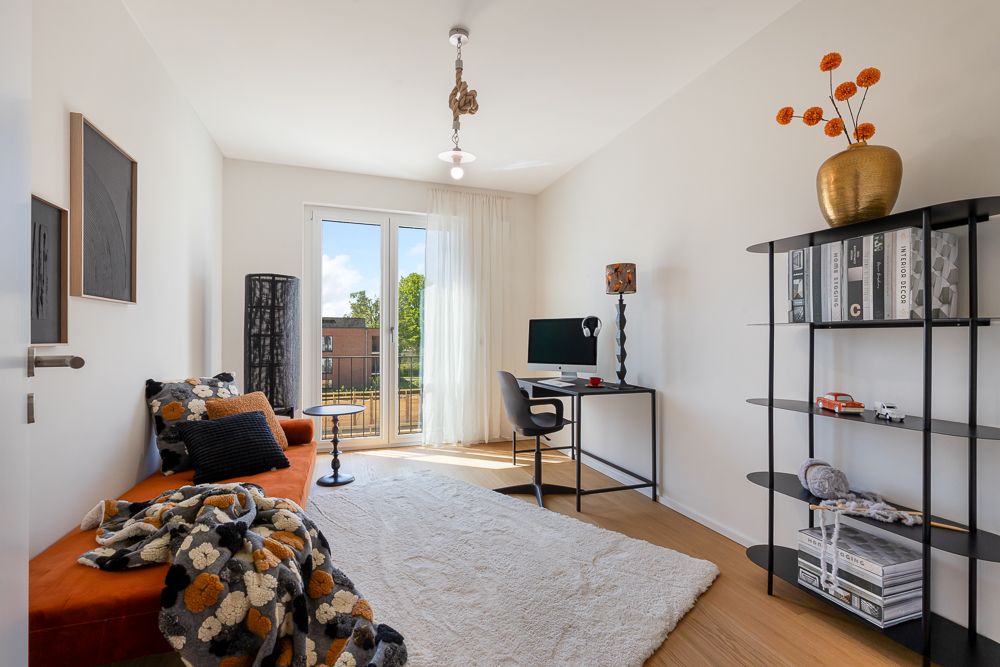 Helles Zimmer mit orangefarbenem Sofa, Schreibtisch, Regal und großem Fenster.