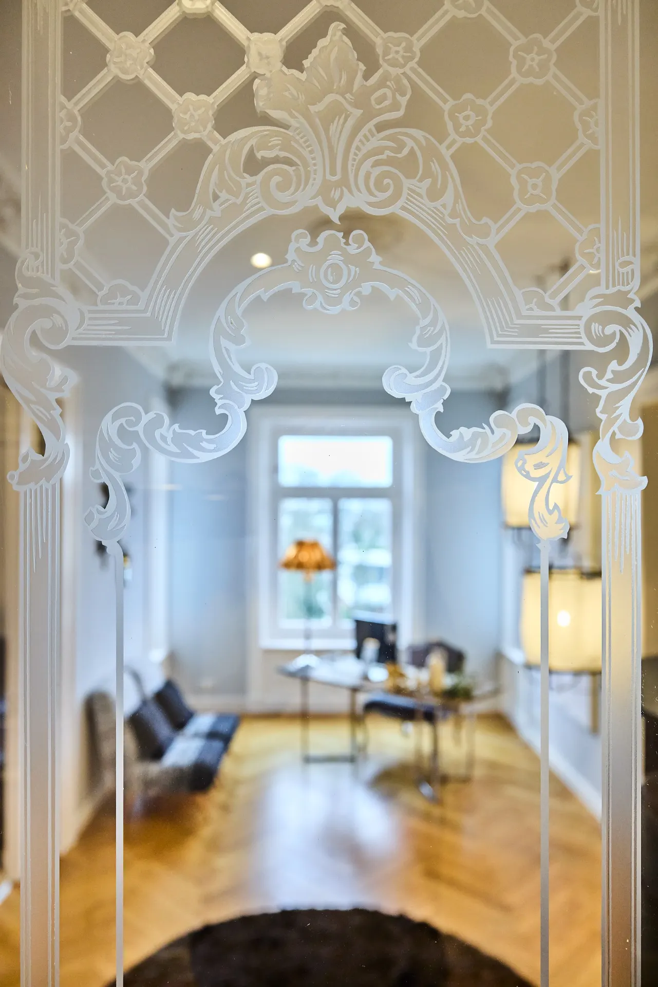 Wohnzimmer mit Parkettboden, Fenster und Glastür mit Ornamentmuster im Vordergrund.