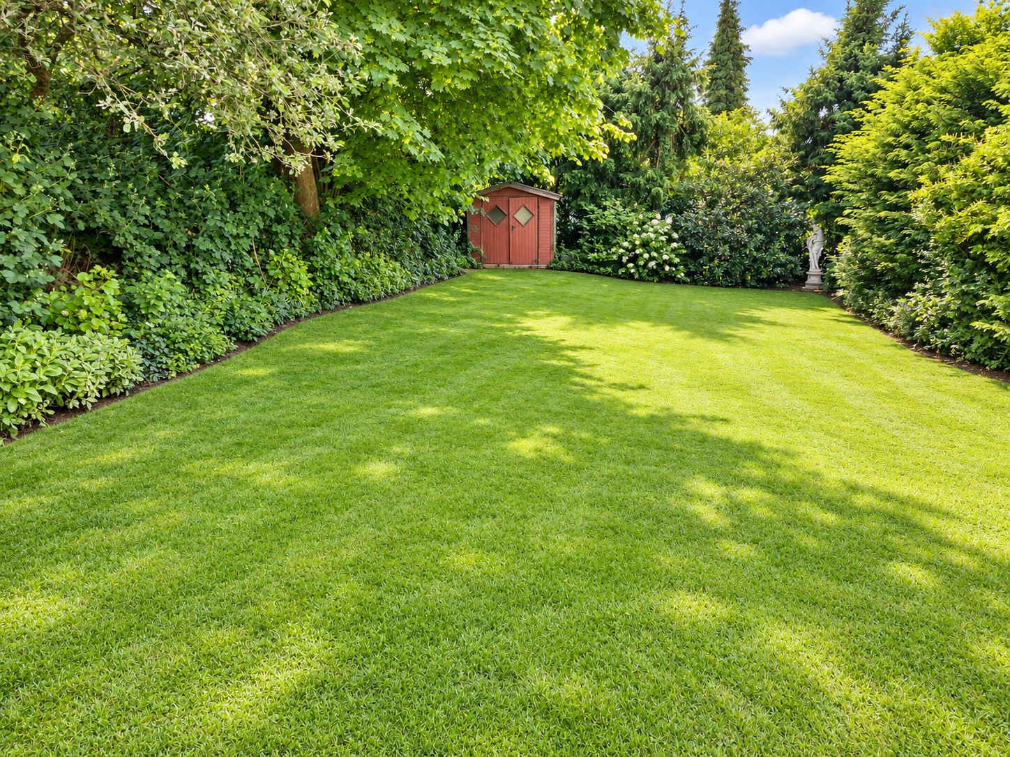 Grüner Garten mit Rasenfläche, umgeben von Büschen und Bäumen, hinten ein kleines rotes Gartenhaus.