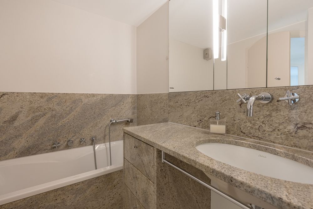 Modernes Badezimmer mit Badewanne, Waschbecken und großem Spiegel, Wände und Ablagen aus Stein.