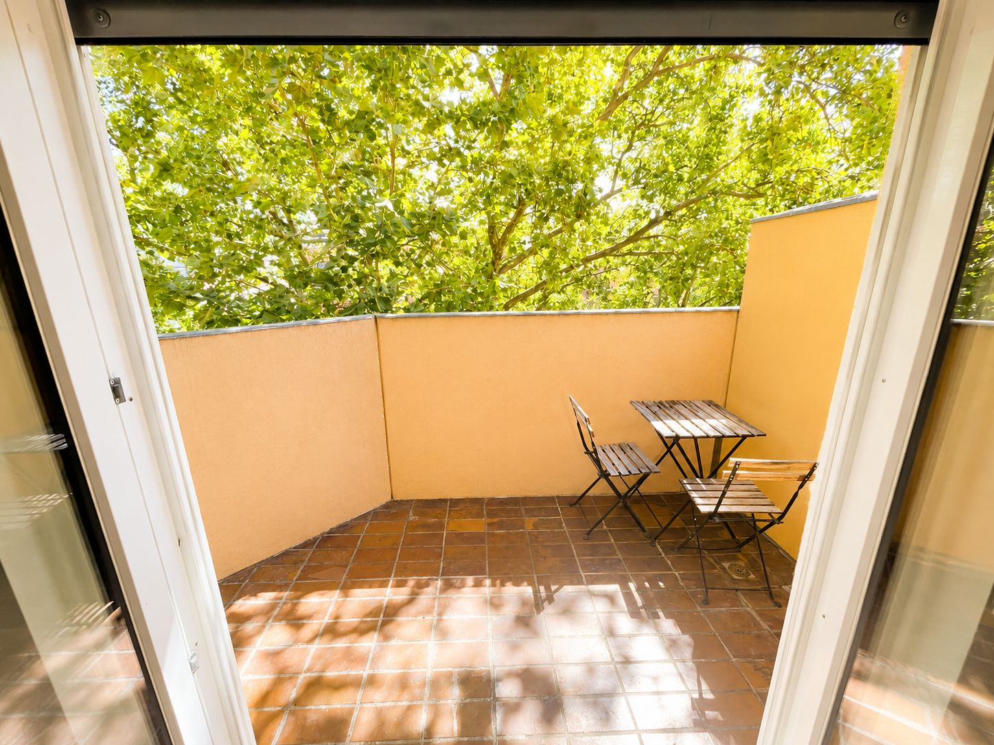 Balkon mit zwei Stühlen und Tisch, Fliesenboden und gelber Wand, im Hintergrund grüne Bäume.
