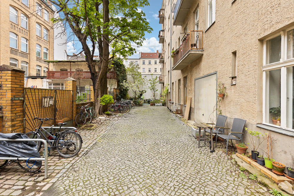 Gepflasterter Innenhof mit Fahrrädern, Bäumen, Stühlen und Blumentöpfen an Wohngebäuden.