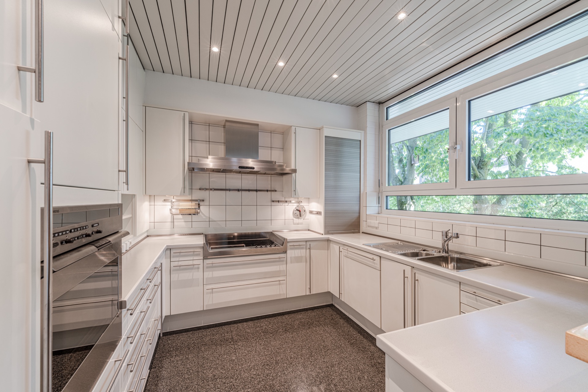 Moderne weiße Einbauküche mit großen Fenstern und Elektrogeräten.