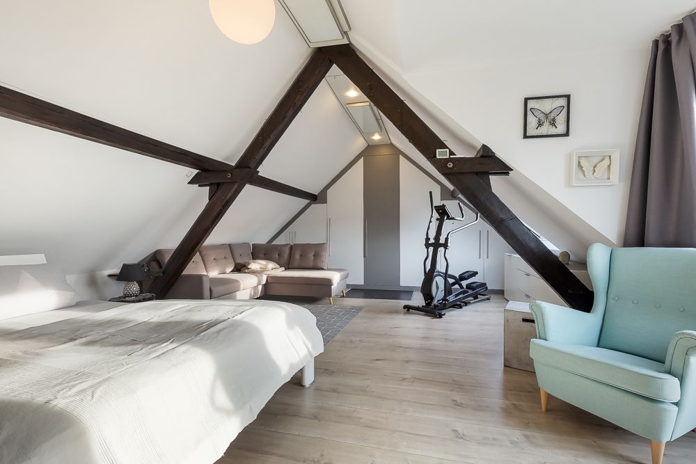 Großes Dachzimmer mit Bett, Sofa, Sessel, Fitnessgerät und sichtbaren Holzbalken.