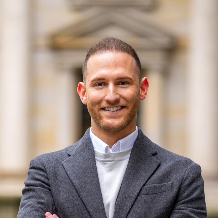 Ein Mann im Anzug steht draußen mit verschränkten Armen und lächelt in die Kamera.