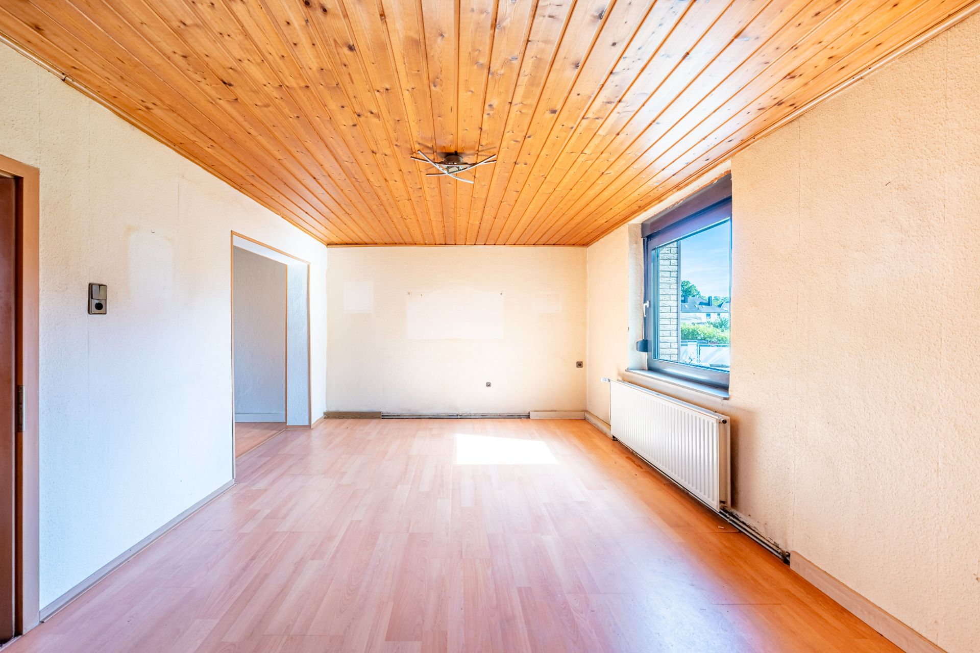 Helles, leeres Zimmer mit Holzdecke, großem Fenster und Heizkörper an der rechten Wand.