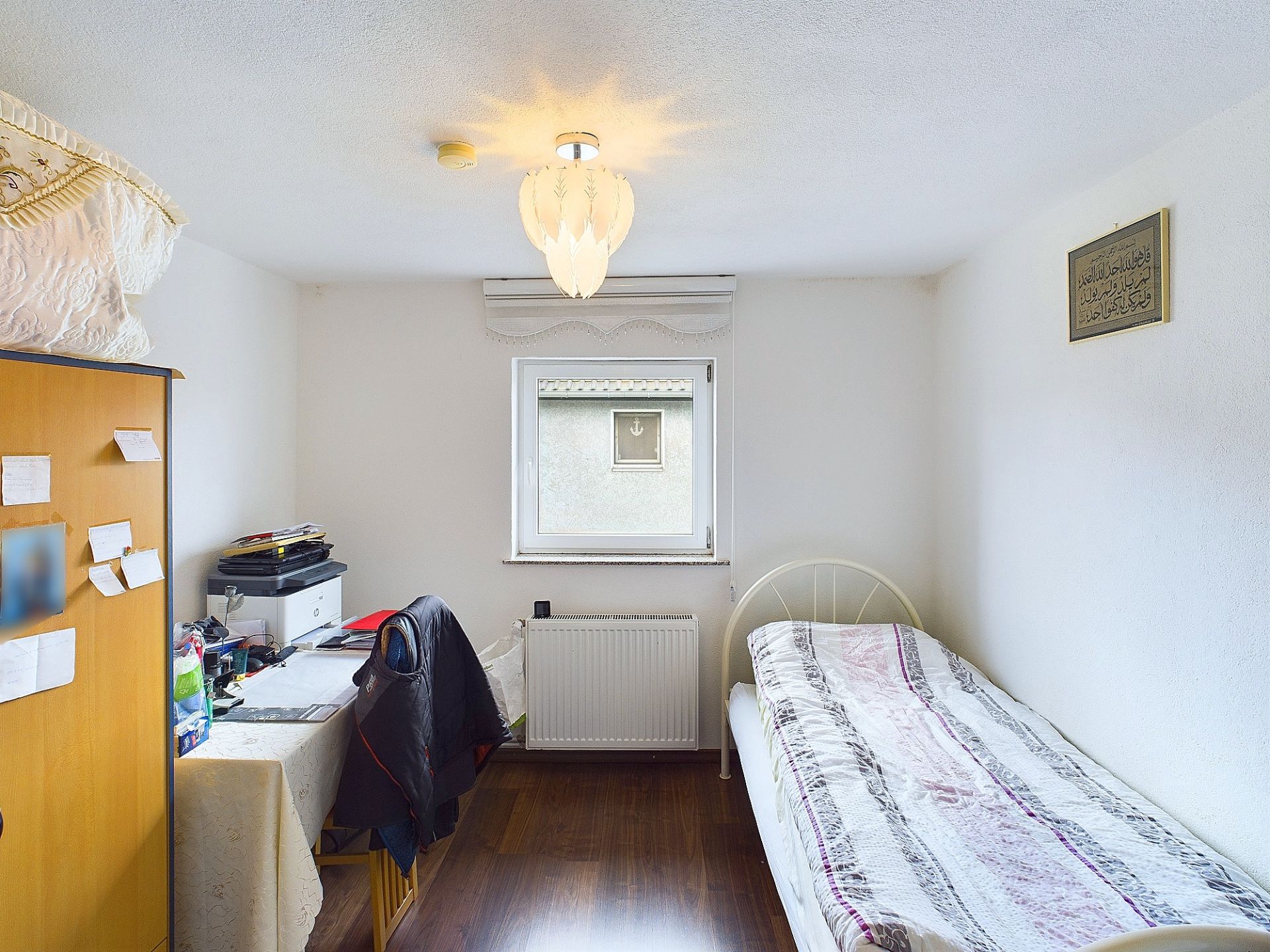 Kleines Zimmer mit Einzelbett, Schreibtisch, Stuhl und Fenster, Holzboden und heller Decke.