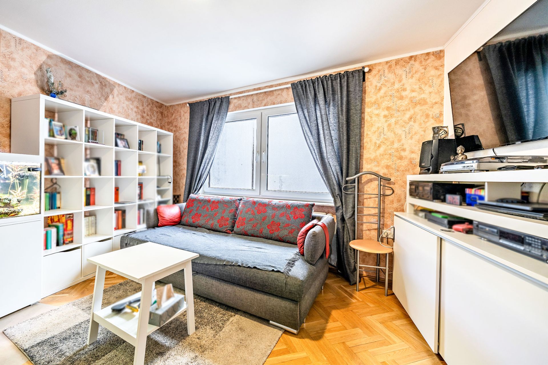 Wohnzimmer mit Sofa, Bücherregal, Couchtisch, TV und großem Fenster mit Vorhängen.