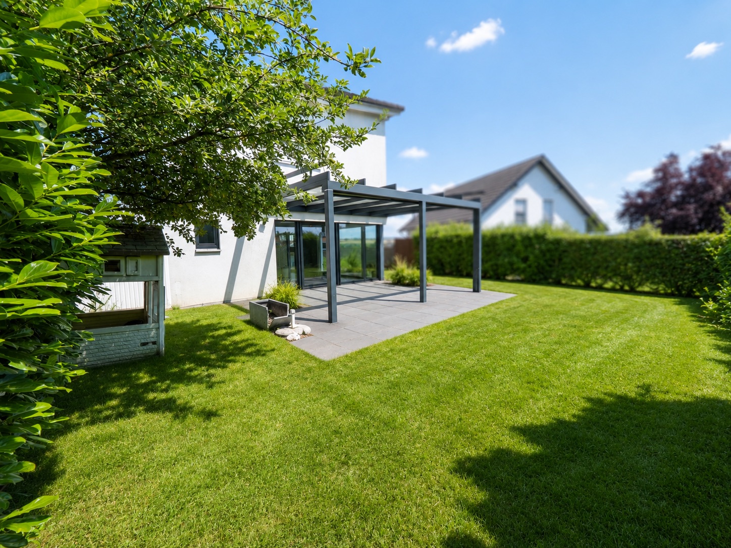 Grüner Garten mit Terrasse und Glasüberdachung an einem weißen Wohnhaus.