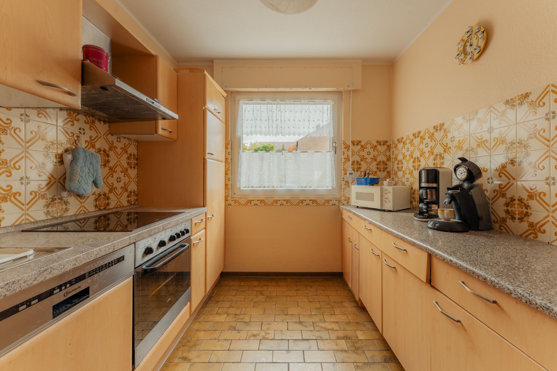 Küche mit Holzschränken, Elektrogeräten und Fenster, gelbem Fliesenboden und gemusterter Wandfliese.