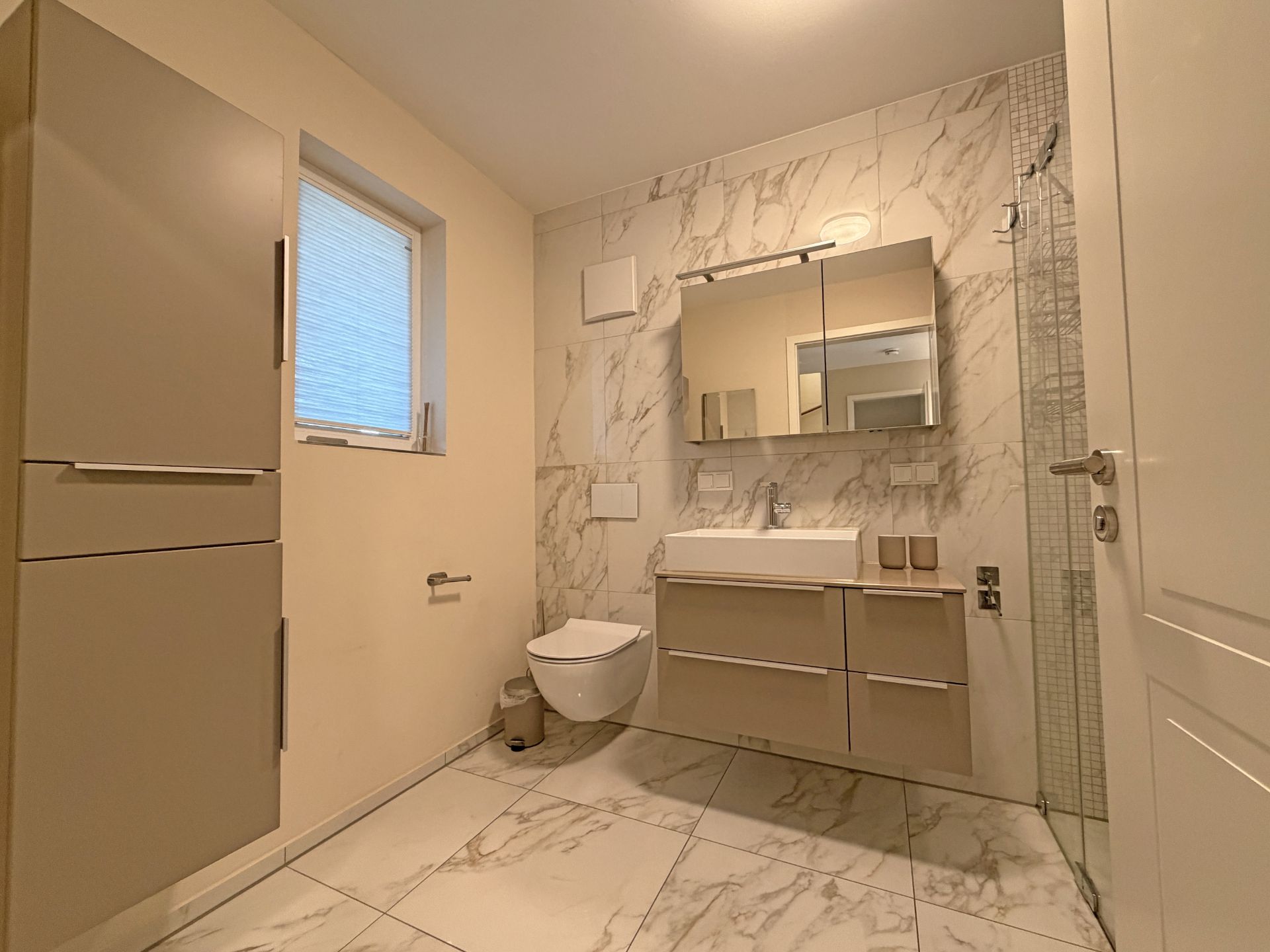 Modernes Badezimmer mit Waschbecken, Spiegel, Hängeschrank, WC und Fenster, Wände in Marmoroptik.
