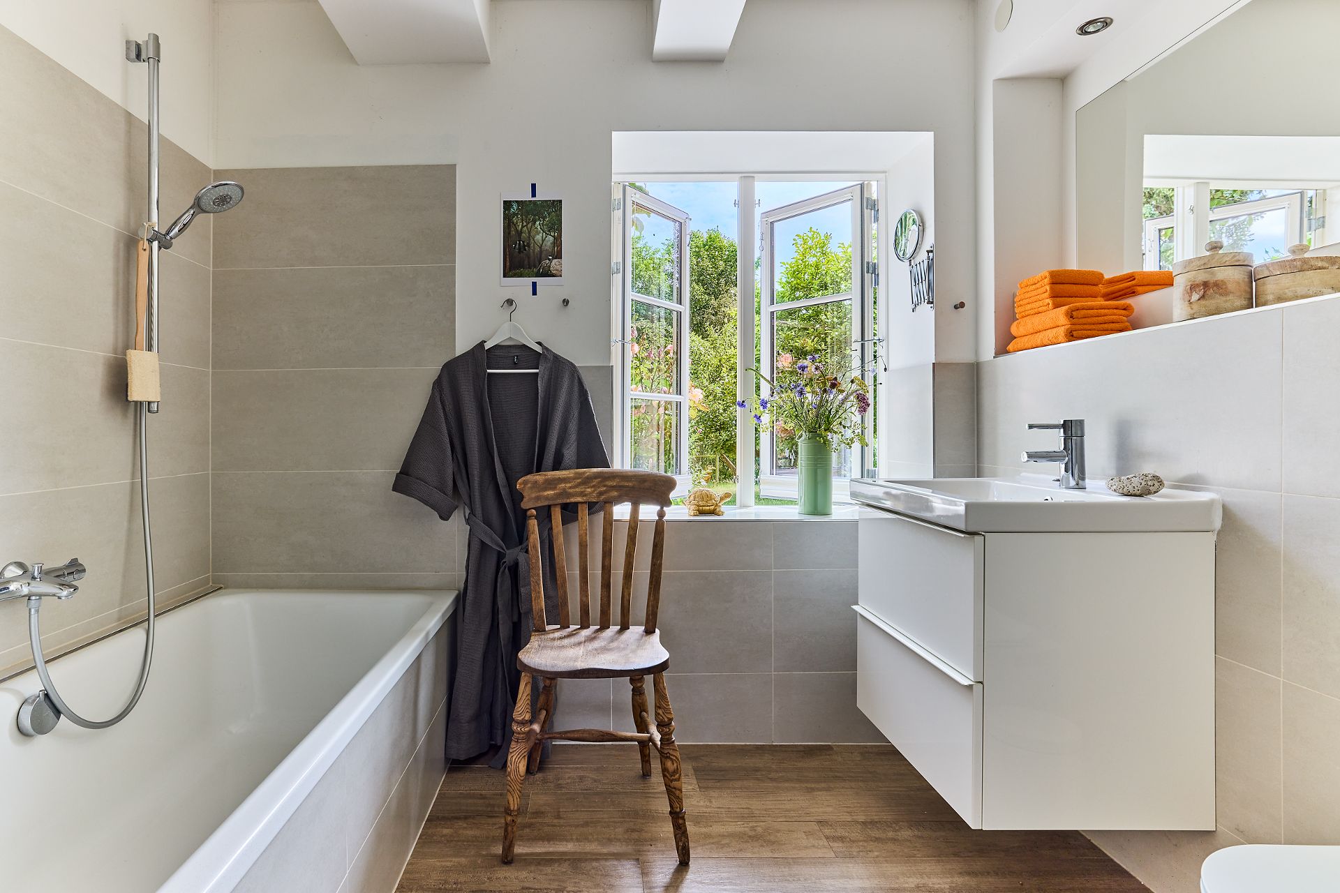 Modernes Badezimmer mit Badewanne, Holzstuhl, Waschbecken und offenem Fenster mit Blick ins Grüne.