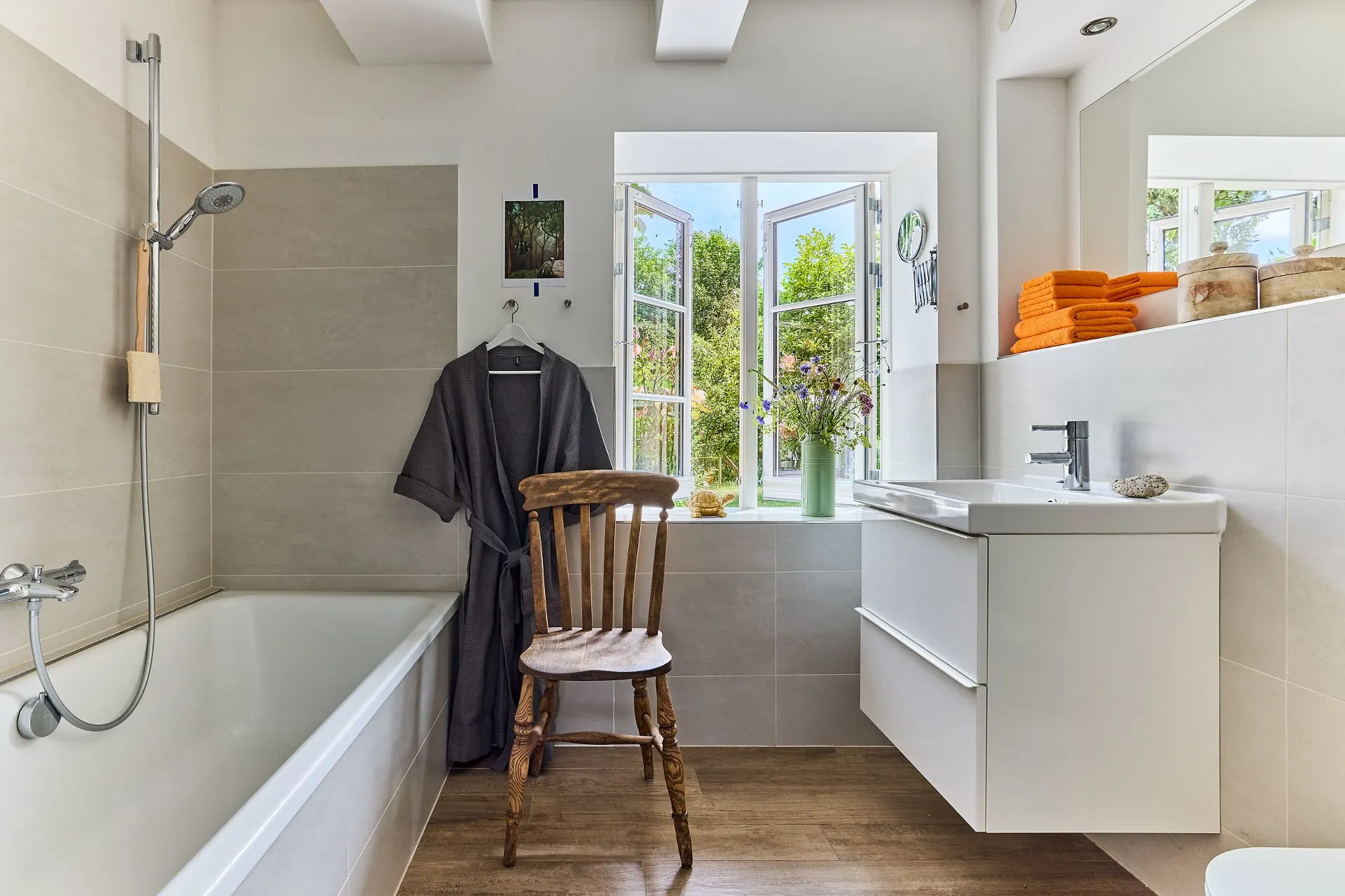 Modernes Badezimmer mit Badewanne, Holzstuhl, Waschbecken und offenem Fenster mit Blick ins Grüne.