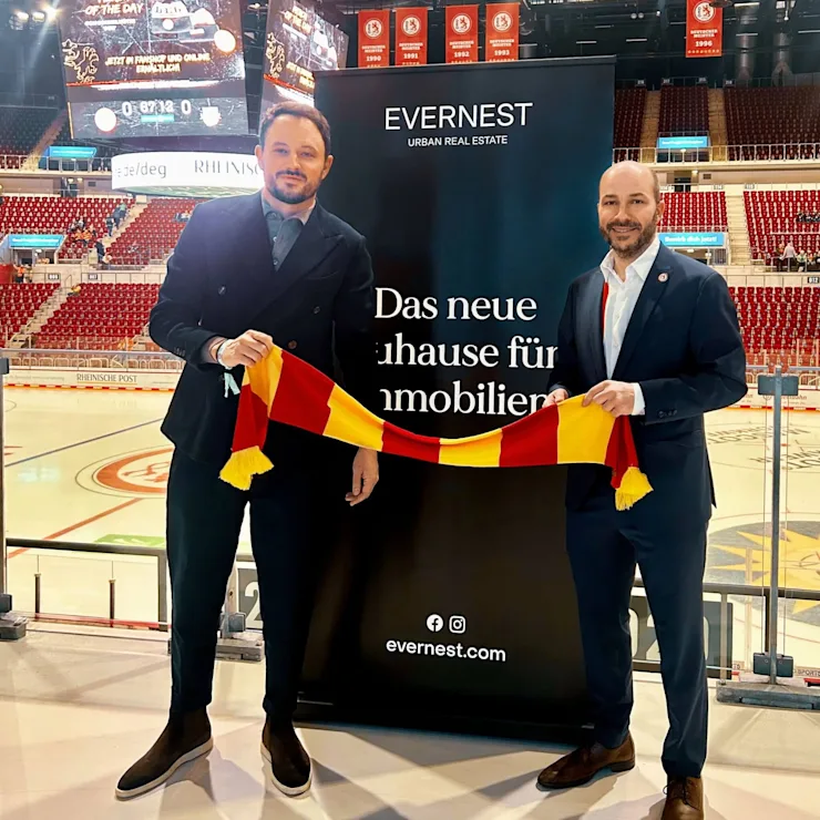 Zwei Männer im Anzug halten einen rot-gelben Schal vor einem Werbebanner in einer Eishalle.