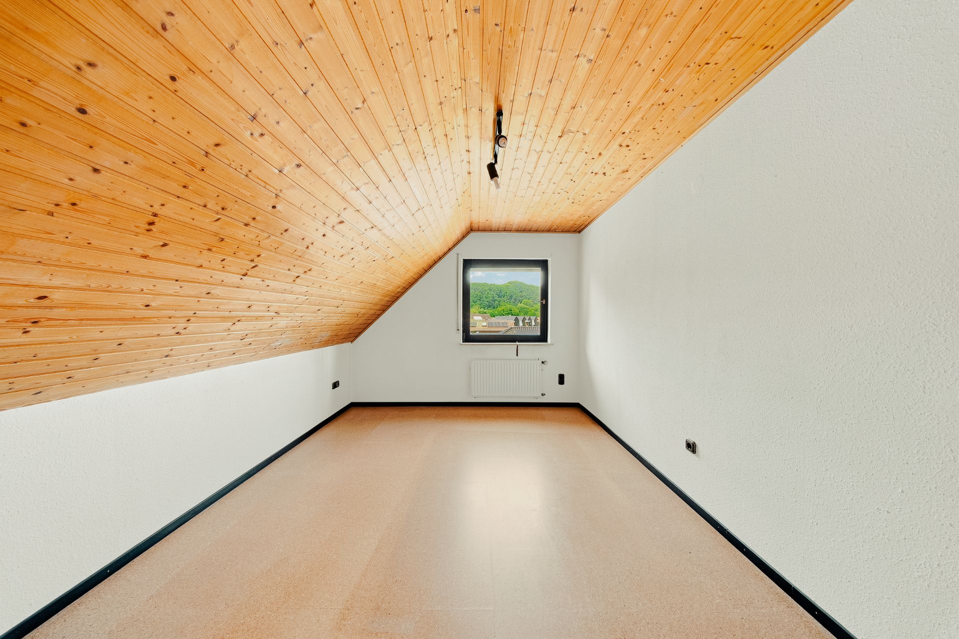 Leerer Raum mit schräger Holzdecke, Fenster und weiß gestrichenen Wänden.