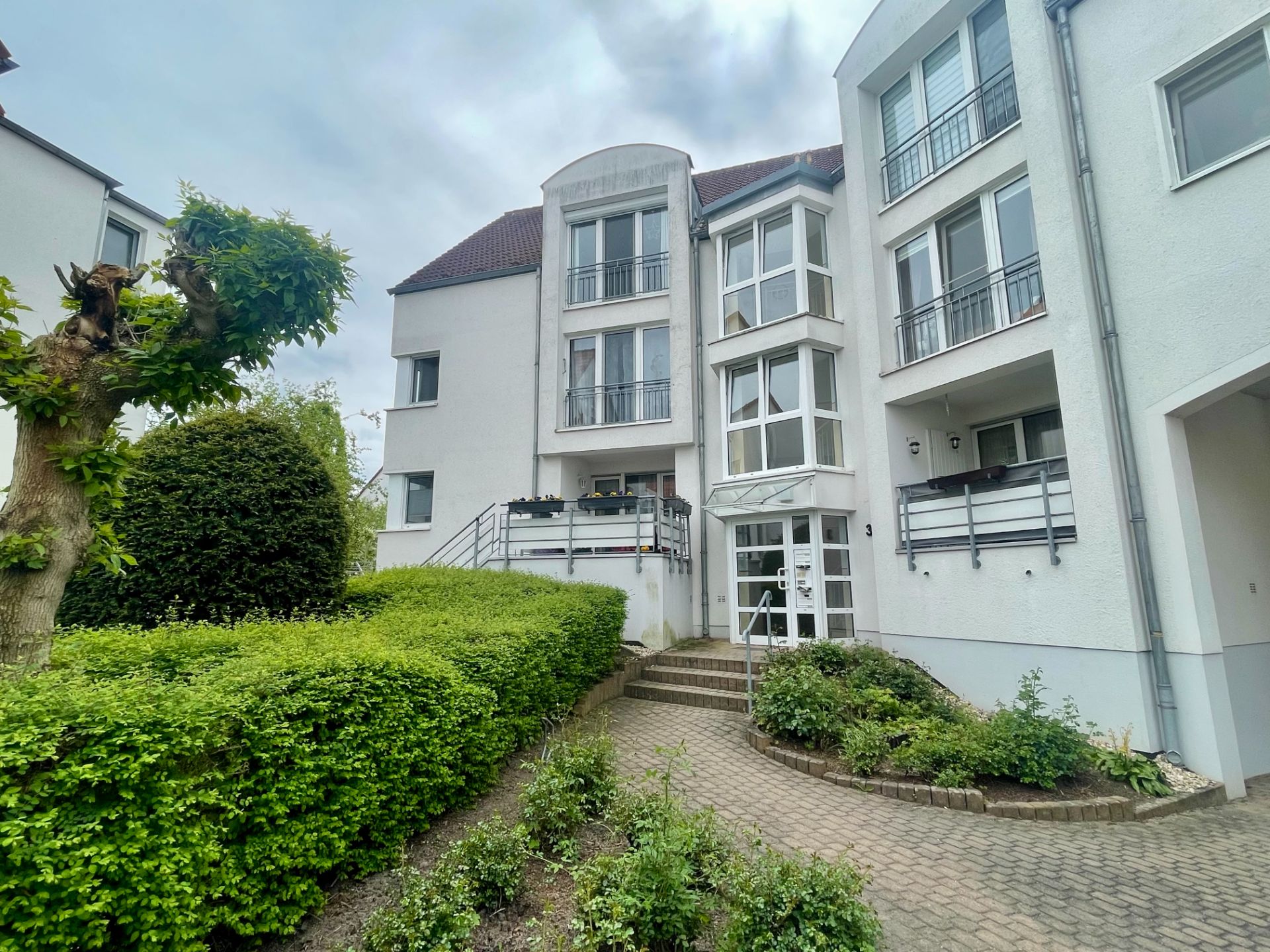 Eingang eines modernen Mehrfamilienhauses mit Balkonen, Treppe und gepflegtem Vorgarten.
