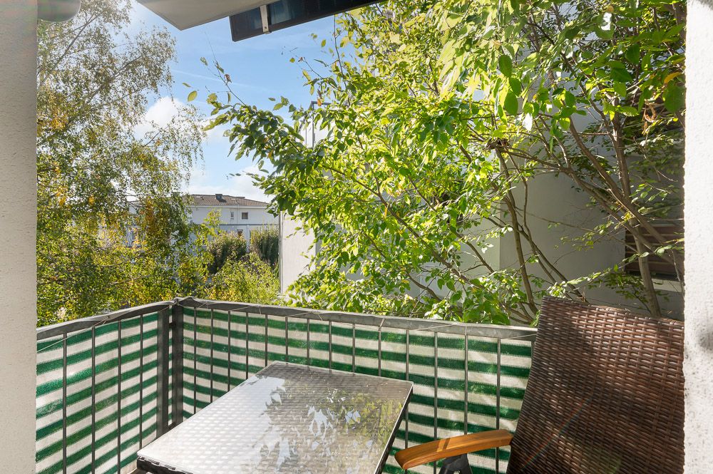 Kleiner Balkon mit Glas­tisch, Stuhl und grünem Sichtschutz, im Hintergrund Bäume.