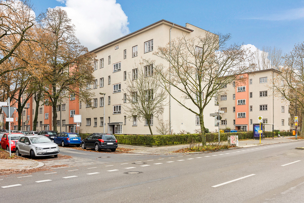 Wohngebäude an einer Straßenecke mit parkenden Autos und Bäumen im Vordergrund.