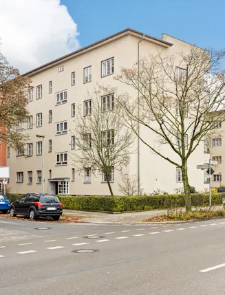 Wohngebäude an einer Straßenecke mit parkenden Autos und Bäumen im Vordergrund.