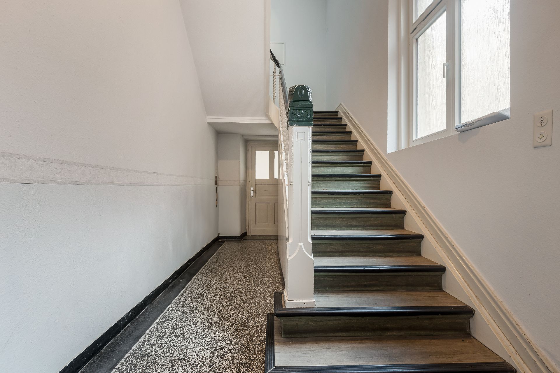 Treppenhaus mit Holztreppe, Fenster an der rechten Wand und Tür am Ende des Flurs.