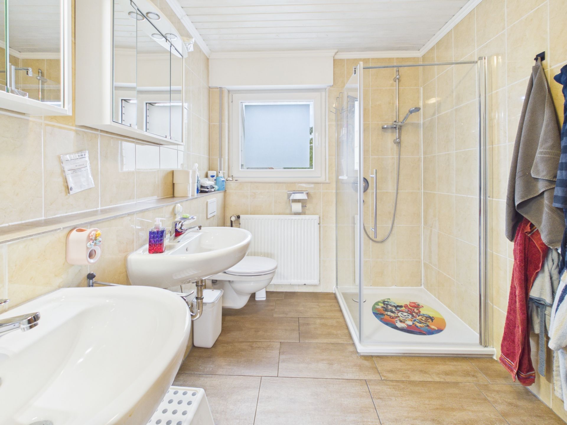 Helles Badezimmer mit zwei Waschbecken, Dusche, WC und Handtüchern an der Wand.