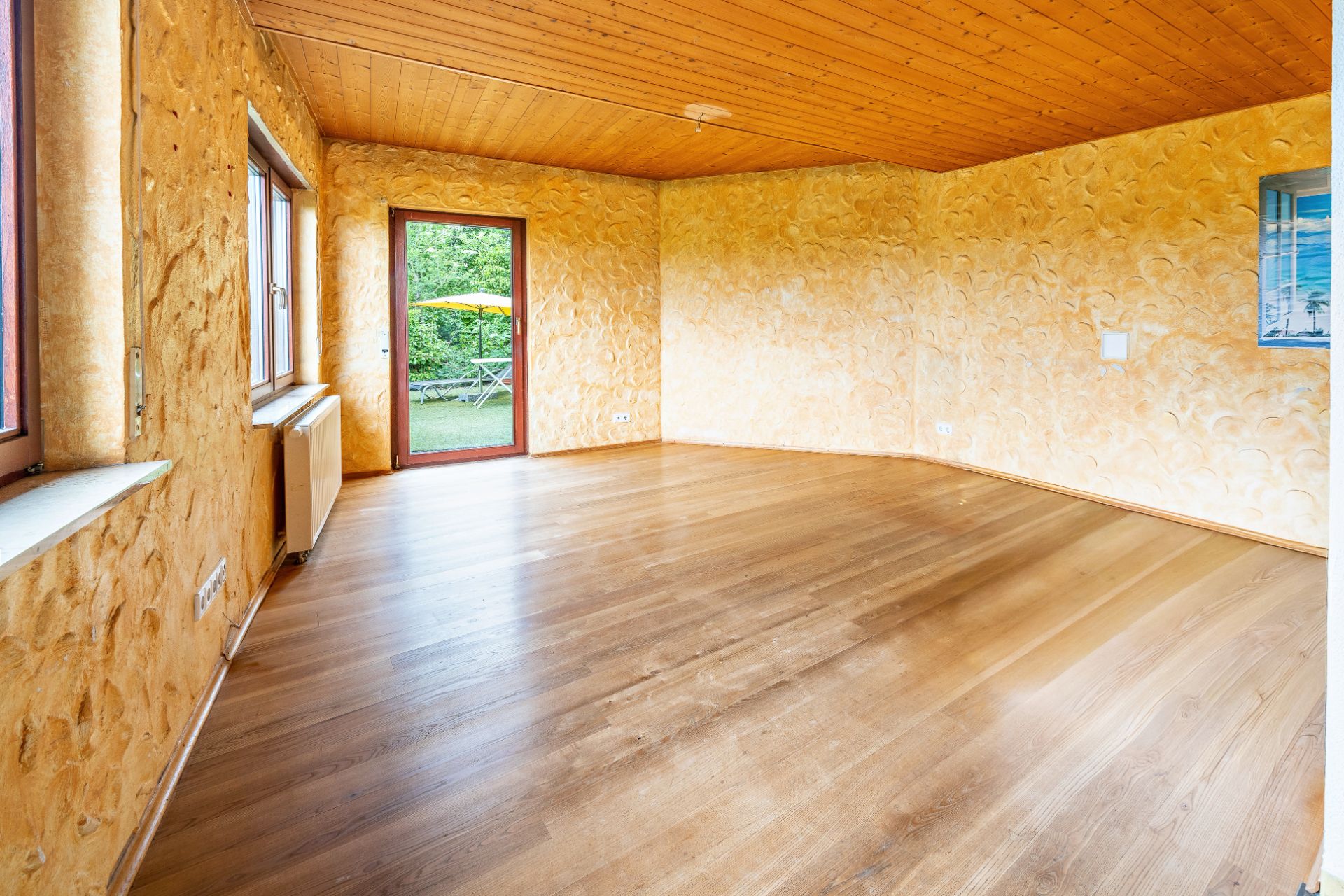 Leeres Zimmer mit Holzboden, gelben Wänden, Fenstern und Tür zum Garten.