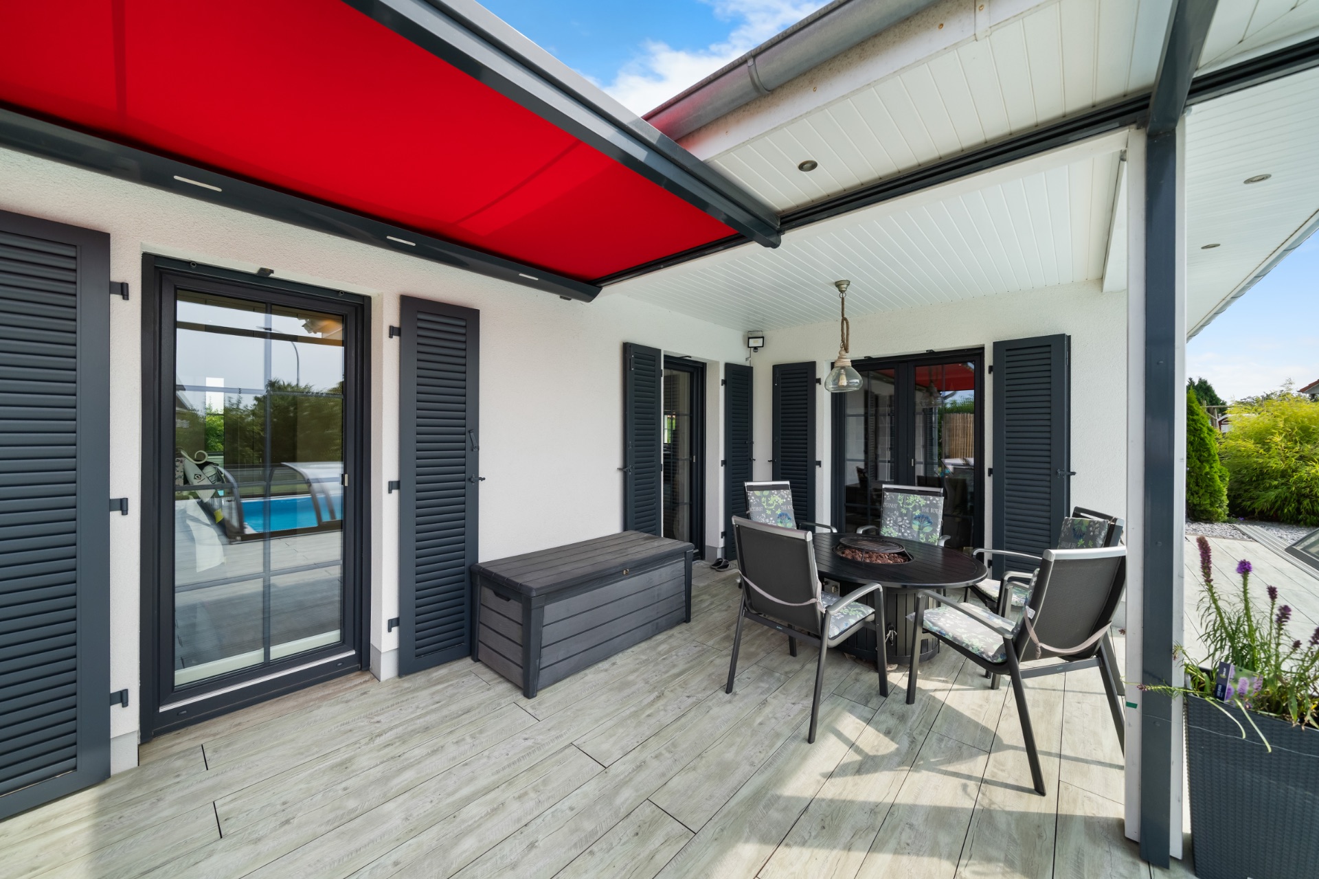 Überdachte Terrasse mit Esstisch, Stühlen und grauen Fensterläden an einem Wohnhaus.