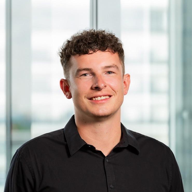 Junger Mann mit lockigen Haaren und schwarzem Hemd steht vor einem großen Fenster.