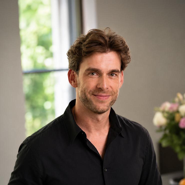 Ein Mann mit dunklem Hemd steht in einem modernen, hellen Raum mit Blumen im Hintergrund.