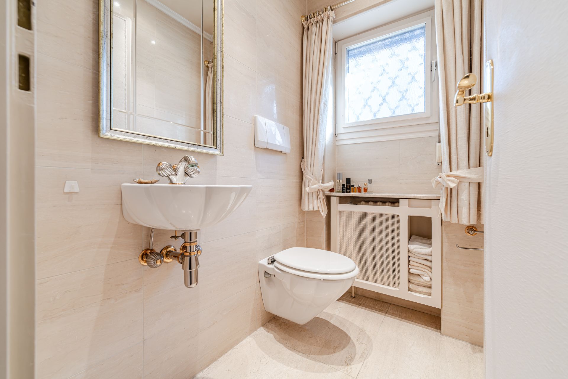 Helles Badezimmer mit Wand-WC, Waschbecken, Spiegel und Fenster mit Vorhängen.