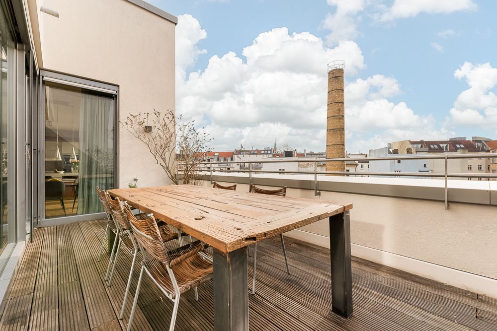 Holztisch mit sechs Stühlen auf einer Dachterrasse, Blick auf Häuser und einen Schornstein.