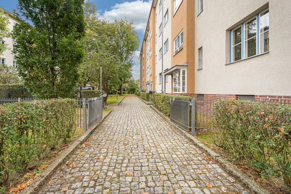 Gepflasterter Weg mit Hecken führt entlang eines Wohnhauses, Bäume im Hintergrund.