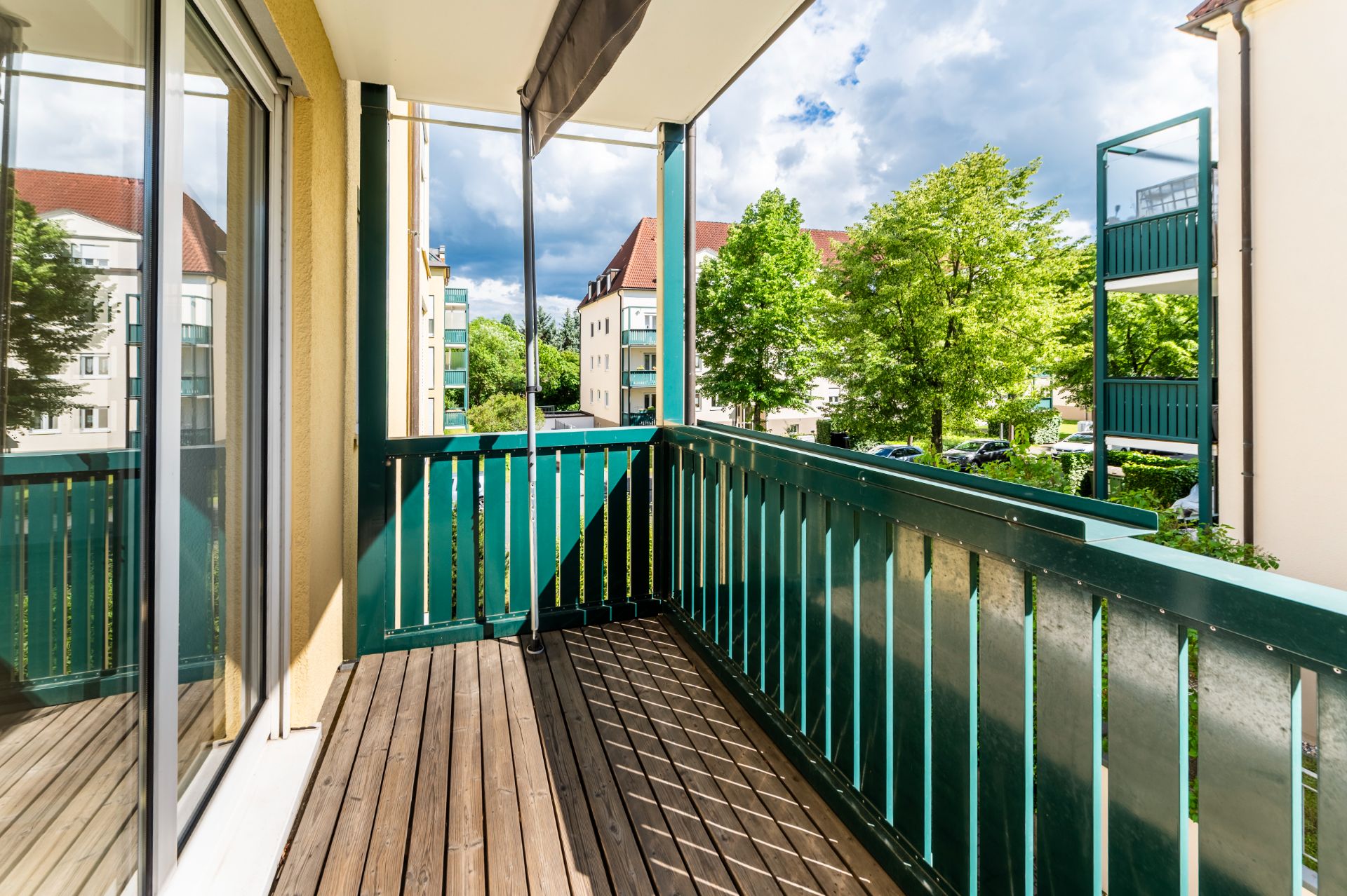 Balkon mit Holzboden und grünem Geländer, Blick auf Bäume und Nachbargebäude.