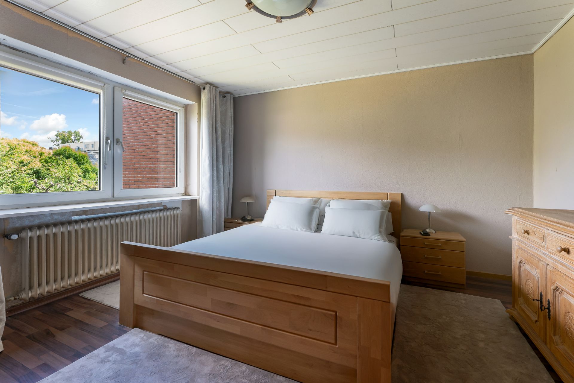 Helles Schlafzimmer mit Holzbett, Nachttischen, Kommode und großem Fenster mit Blick ins Grüne.