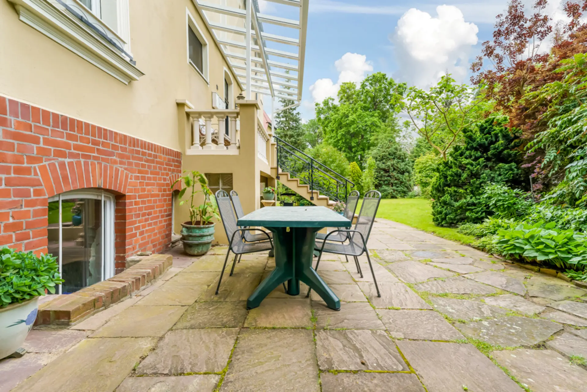Steinterrasse mit Tisch, sechs Stühlen und Pflanzen neben einem Wohnhaus und Garten.