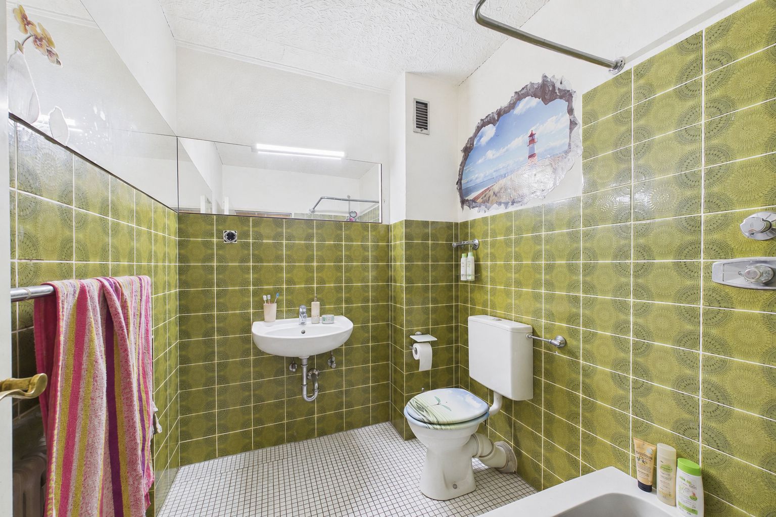 Badezimmer mit grünen Fliesen, Waschbecken, Toilette, Handtüchern und Wandbild eines Leuchtturms.