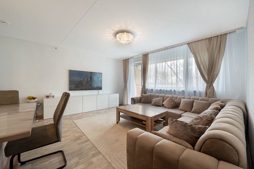 Helles Wohnzimmer mit großer Eckcouch, Couchtisch, Esstisch, TV und bodentiefen Fenstern mit Vorhängen.